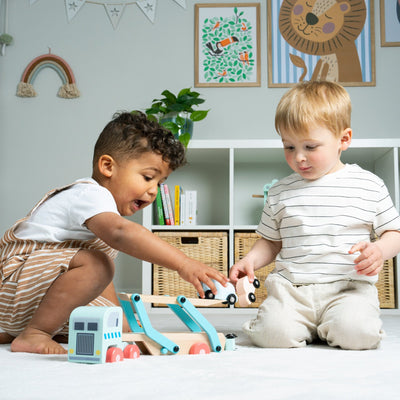 Two young children sit on the floor playing with the TP Toys Owl & Fox Wooden Vehicle Transporter – FSC® certified, in a bright room decorated with art and storage baskets.