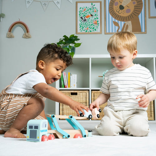 Two young children sit on the floor playing with the TP Toys Owl & Fox Wooden Vehicle Transporter – FSC® certified, in a bright room decorated with art and storage baskets.