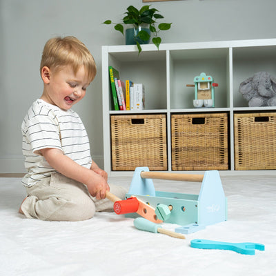 A young child sits on a white carpet playing with the TP Toys Owl & Fox Wooden Workbench Set with Tool Caddy – FSC® certified, enjoying imaginative pretend play, with a shelf and baskets in the background.