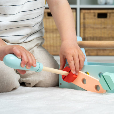 A child uses a wooden screwdriver from the TP Toys Owl & Fox Wooden Tool Caddy - FSC® certified to insert a red screw into a pastel activity board, encouraging imaginative play on a carpeted floor.