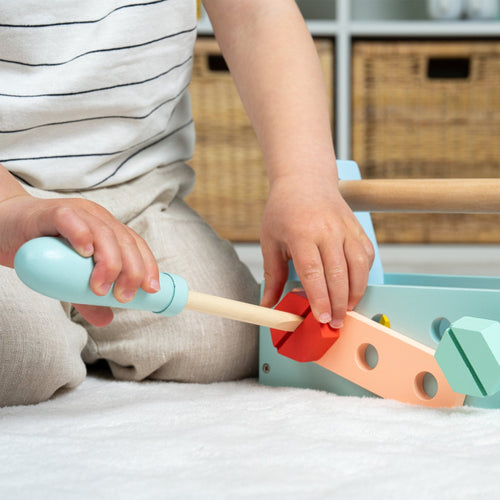A child uses a wooden screwdriver from the TP Toys Owl & Fox Wooden Tool Caddy - FSC® certified to insert a red screw into a pastel activity board, encouraging imaginative play on a carpeted floor.