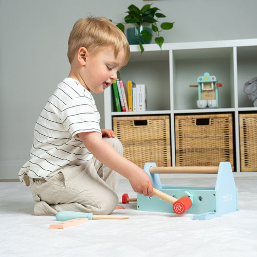 A young child plays imaginatively on a carpeted floor with the TP Toys Owl & Fox Wooden Tool Caddy (FSC® certified), surrounded by shelves and baskets.