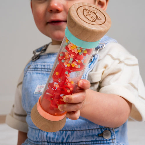 A baby in denim overalls holds the TP Toys Owl & Fox Wooden Rainmaker - FSC® certified, a rattle filled with colorful beads thats perfect for sensory development.
