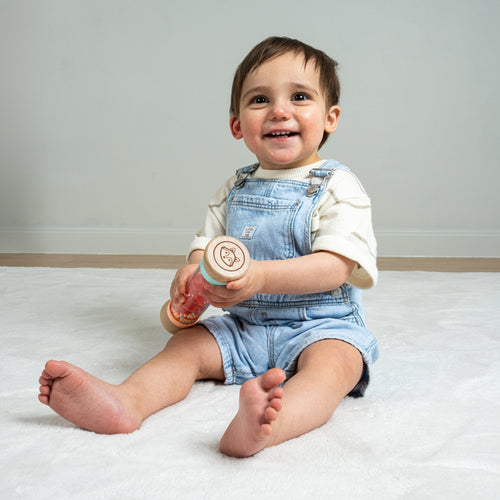 A smiling toddler in denim overalls sits on a white carpet, holding the TP Toys Owl & Fox Wooden Rainmaker - FSC® certified, a musical toy that enhances sensory development.