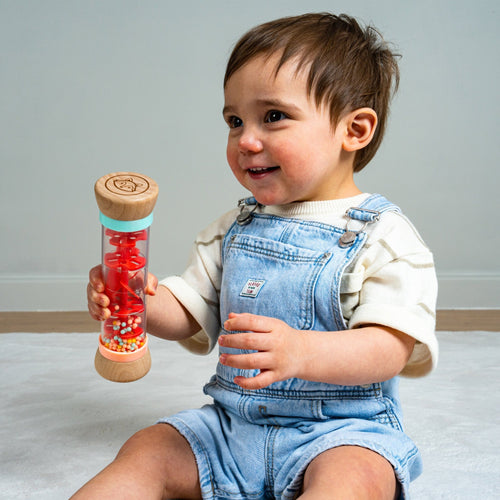 A toddler in denim overalls sits on the floor, smiling and holding the TP Toys Owl & Fox Wooden Rainmaker—an FSC® certified musical toy filled with colorful beads, perfect for sensory development.