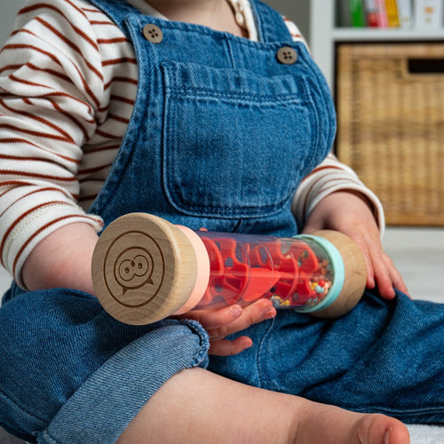 A seated child in blue denim overalls holds the TP Toys Owl & Fox Wooden Rainmaker—an FSC®-certified clear musical toy with wooden ends and colorful beads, ideal for sensory development.