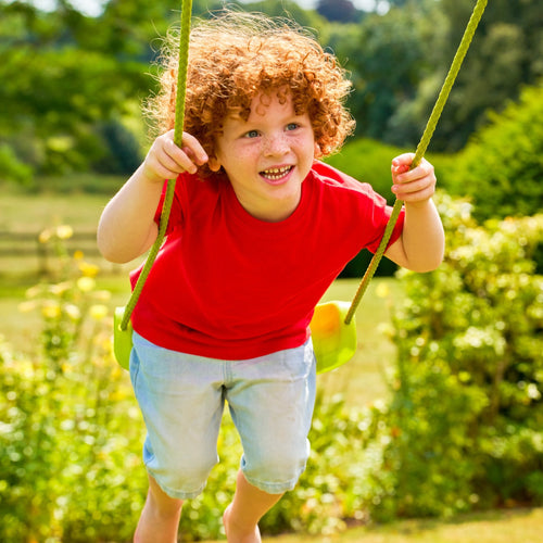 A young child with curly red hair, in a red shirt and light shorts, smiles while swinging outdoors on the TP Toys TP Metal Swing Slide & Glide playset in a sunny green garden.