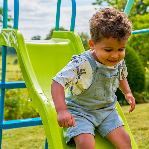 A young child enjoys a sunny day outdoors, sliding down the bright green slide of the TP Metal Swing Slide & Glide garden playset by TP Toys.