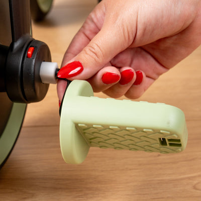 A hand with red-painted nails attaches or detaches a green textured pedal to a black mechanism on the TP Toys Zoomee Balance Bike to Trike, shown on a wooden surface.