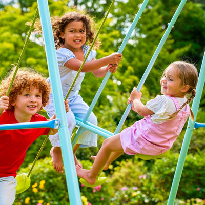 Three young children are smiling and playing together on the TP Toys TP Metal Swing Slide & Glide in a green, sunny park.