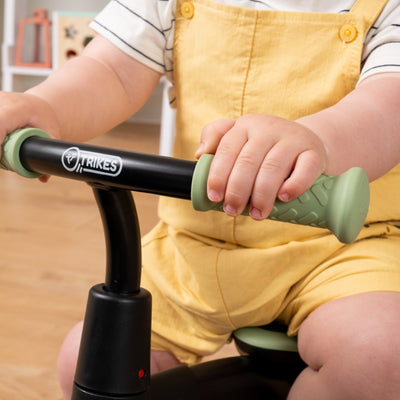 A young child rides the Zoomee Balance Bike to Trike by TP Toys indoors, gripping the green handles of the black trike to build confidence and support development.