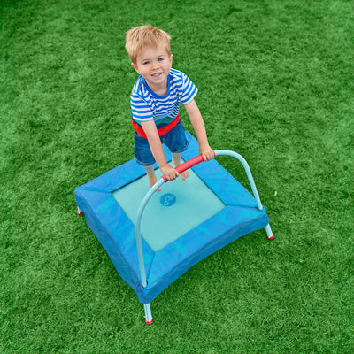 A young child in a striped shirt smiles up at the camera while standing on the TP Toys TP Junior Trampoline with a handlebar, set on green grass.