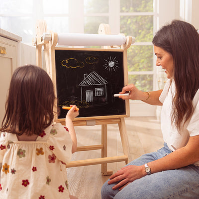 An adult and child use the TP Toys TP Active-Tots Pikler Style Wooden Easel Play Accessory (FSC® certified) to draw a house, sun, and clouds on the chalkboard and paper scroll in a bright room.
