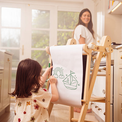 A young child draws on a TP Toys TP Active-Tots Pikler Style Wooden Easel Play Accessory – FSC® certified, with a large paper roll in the kitchen, while an adult woman smiles nearby.