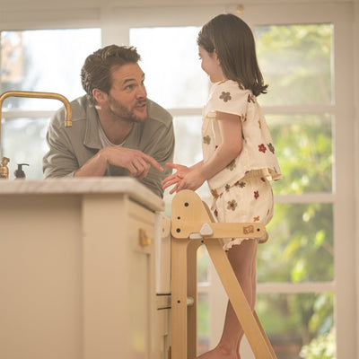 An adult talks to a young girl standing on the TP Toys TP Active-Tots Pikler Style Wooden Folding Helper Tower With Play Easel & Play Mats—FSC® certified—by the kitchen counter, sunlight streaming through large windows.