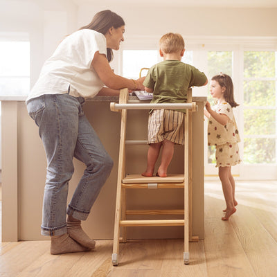 In a bright kitchen, an adult and two children interact. One child stands at the counter on the TP Toys Active-Tots Pikler Style Wooden Folding Helper Tower With Play Easel & Play Mats (FSC® certified), while the other holds a cup nearby.