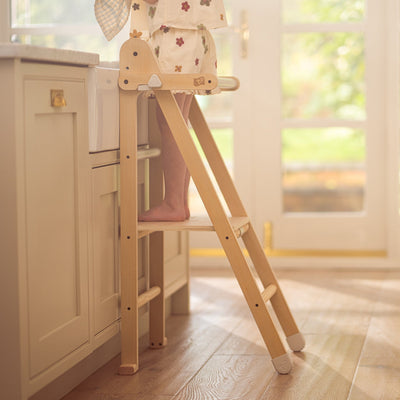 A child stands on the TP Toys TP Active-Tots Pikler Style Wooden Folding Helper Tower with Play Easel & Play Mats—FSC® certified—at a kitchen counter near a sink, sunlight streaming in through the glass doors behind.