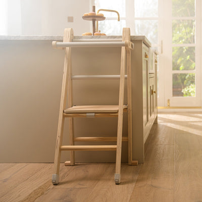 The TP Active-Tots Pikler Style Wooden Folding Helper Tower With Play Easel & Play Mats by TP Toys is placed by a kitchen island with pastries in the background, as sunlight pours through glass doors for a warm, inviting scene.