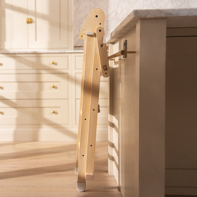 The TP Active-Tots Pikler Style Wooden Folding Helper Tower With Play Easel & Play Mats by TP Toys is shown folded and leaning against a kitchen counter, positioned between the counter and the cabinet wall.