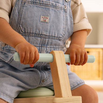 A toddler in denim overalls grips the handlebar of a TP Toys TP Active-Tots Pikler Style Toddler Bouncer - FSC® certified while playing indoors.