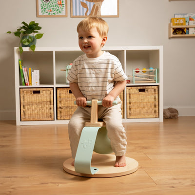 A young child sits barefoot on the TP Active-Tots Pikler Style Toddler Bouncer by TP Toys, FSC® certified, in a tidy, well-lit playroom with shelves and baskets in the background.