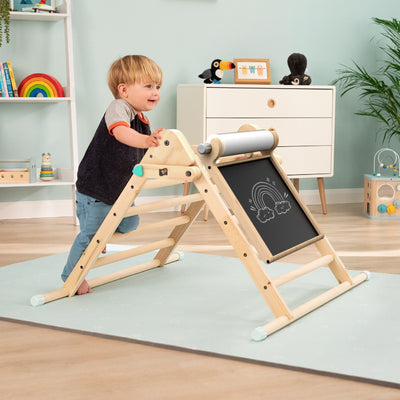 A young child stands by the TP Toys TP Active-Tots Pikler Style Wooden Climbing Triangle With Wooden Play Easel—an FSC® certified indoor Montessori toy—inside a playroom decorated with toys and wall art.
