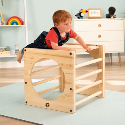 A young child climbs on the TP Toys TP Active-Tots Pikler Style Wooden Climbing Cube With Play Easel (FSC® certified) in a bright, tidy indoor play area with toys and shelves in the background.