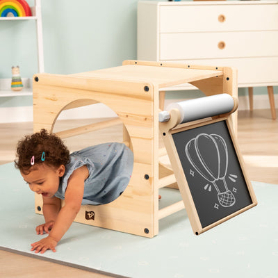 A toddler explores the TP Active-Tots Pikler Style Wooden Climbing Cube With Play Easel by TP Toys, featuring a chalkboard easel on the side, creating an engaging and eco-friendly indoor play experience.