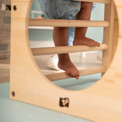 A toddler climbs barefoot on the TP Toys Active-Tots Pikler Style Wooden Climbing Cube With Play Easel—FSC® certified—in an indoor play area.