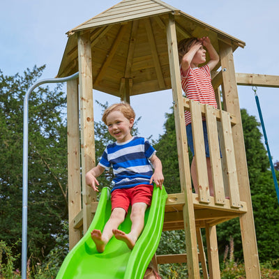 Two young boys play on the TP Toys Castlewood climbing frame with Double Wooden Swing Arm & 1.8m Wavy Slide (FSC® certified); one slides down the wavy slide while the other stands in the upper play tower, looking out.