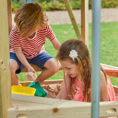 Two children play in a wooden sandbox outdoors. Nearby, the TP Toys Castlewood climbing frame with Double Wooden Swing Arm & 1.8m Wavy Slide (FSC® certified) offers more adventure as one plays with sand and the other digs with her hands.