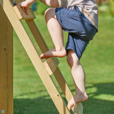 A barefoot child in blue shorts climbs the TP Toys Castlewood climbing frame with Double Wooden Swing Arm & 1.8m Wavy Slide (FSC® certified) outdoors on a sunny day.