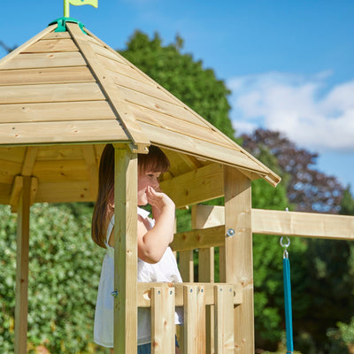 A young girl stands in a TP Toys Castlewood climbing frame with Double Wooden Swing Arm & 1.8m Wavy Slide (FSC® certified), outdoors, looking out with her hand near her mouth. Trees and blue sky are visible behind her.