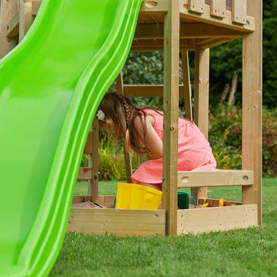 A young girl in a pink dress plays in a sandbox beneath the TP Toys Castlewood climbing frame with Double Wooden Swing Arm and 1.8m wavy slide—FSC® certified—set up in a grassy yard.