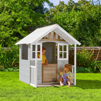 Two children play in the TP Toys Snowdrop Cottage Wooden Playhouse—an FSC® certified wooden playhouse with white trim—set on green grass among trees and a brick wall in the background.