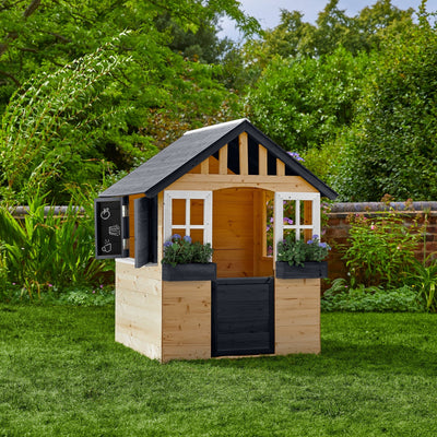 The TP Toys TP Blackberry Cottage Wooden Playhouse—FSC® certified—features white windows, black trim, flower boxes, and a chalkboard sign. Set on grass among greenery, its perfect for imaginative play.