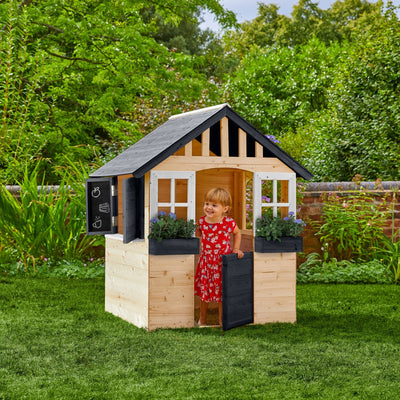 A young child in a red floral dress stands at the open door of the TP Toys TP Blackberry Cottage Wooden Playhouse – FSC® certified, set in a grassy backyard with flower boxes.