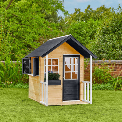 The TP Toys Bluebell Cottage Wooden Playhouse, FSC® certified, features a black roof and door, white trim, and a window box—an ideal garden playhouse for imaginative fun set against greenery and a brick wall.