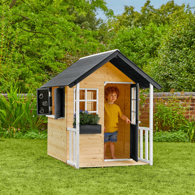 A young child stands in the doorway of the TP Toys Bluebell Cottage Wooden Playhouse – FSC® certified – with black roof panels, ready for imaginative play in a grassy backyard.