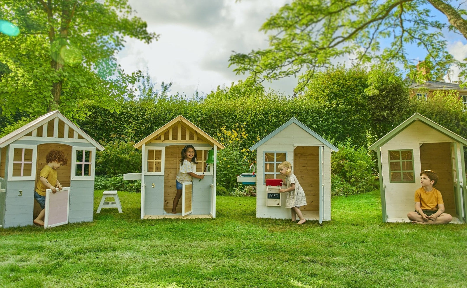 Four children enjoy imaginative play in separate TP Toys Lavender Cottage Wooden Playhouses, made from FSC® certified wood, set on a grassy lawn surrounded by trees and greenery on a bright day.
