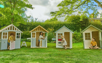 Four children enjoy imaginative play in separate TP Toys Lavender Cottage Wooden Playhouses, made from FSC® certified wood, set on a grassy lawn surrounded by trees and greenery on a bright day.