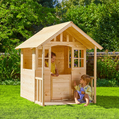Two children enjoy imaginative play in and around the Clover Cottage Wooden Playhouse by TP Toys, FSC® certified, set on a grassy lawn surrounded by trees and shrubs.