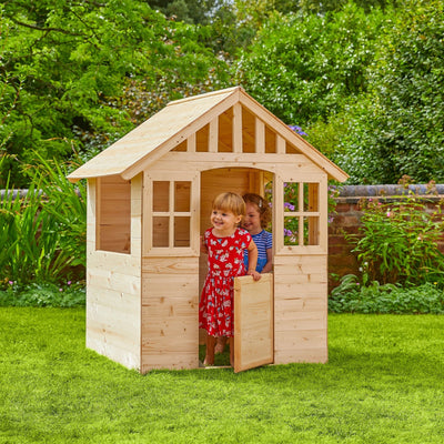 Two young children enjoy imaginative play in the Dandelion Cottage Wooden Playhouse by TP Toys, crafted from FSC® certified wood. Set on a grassy lawn with trees and a brick wall behind, it promises endless outdoor fun.