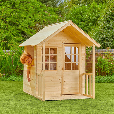 Two children enjoy imaginative play in and around the TP Toys Lavender Cottage Wooden Playhouse—crafted from FSC® certified wood—set amidst a grassy garden with lush plants and trees.