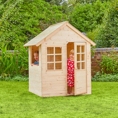 Two children enjoy imaginative play in and around the TP Toys Foxglove Cottage Wooden Playhouse, made from FSC® certified wood, set on a grassy lawn with trees and bushes in the background.