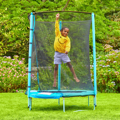 A child in a yellow sweatshirt jumps on a TP Toys UP 4.5ft Junior Trampoline with a zip-free IGLOO door, set up on green grass among flowering bushes in the garden.