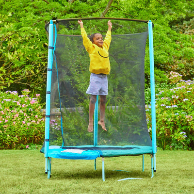 A child in a yellow sweatshirt jumps on a TP Toys UP 4.5ft Junior Trampoline with a zip-free IGLOO door and safety net, set up in a grassy backyard surrounded by greenery and flowers.