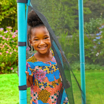 A young girl in a colorful dress smiles while standing at the entrance of a TP Toys TP UP 4.5ft Junior Trampoline, featuring a zip-free IGLOO door, set in a charming garden.