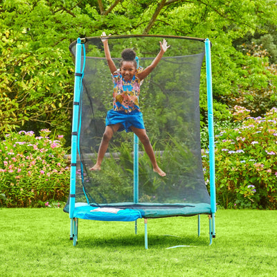 A child jumps with arms raised on a TP Toys UP 4.5ft Junior Trampoline with safety netting in a garden filled with green grass and flowering plants.