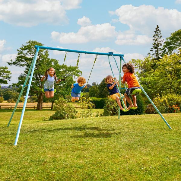 Four children play on the TP Toys TP Triple Metal Swing Set with Glider Swing Seat, which features adjustable seats. The blue swing set stands on grassy park grounds, surrounded by trees and greenery on a sunny day.